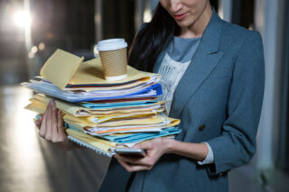 Businesswoman carrying stack of file folders and coffee while using cell phone